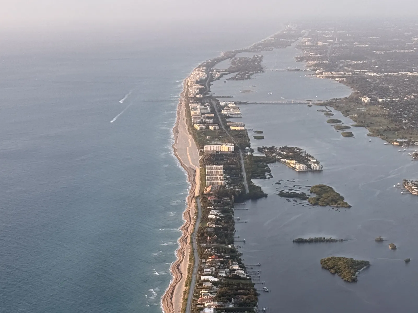 Hazy high aerial view down the long narrow barrier island: a thin line of beach and buildings between the ocean on the left and the lagoon on the right, with small islands, a bridge and the mainland shore stretching to the horizon.