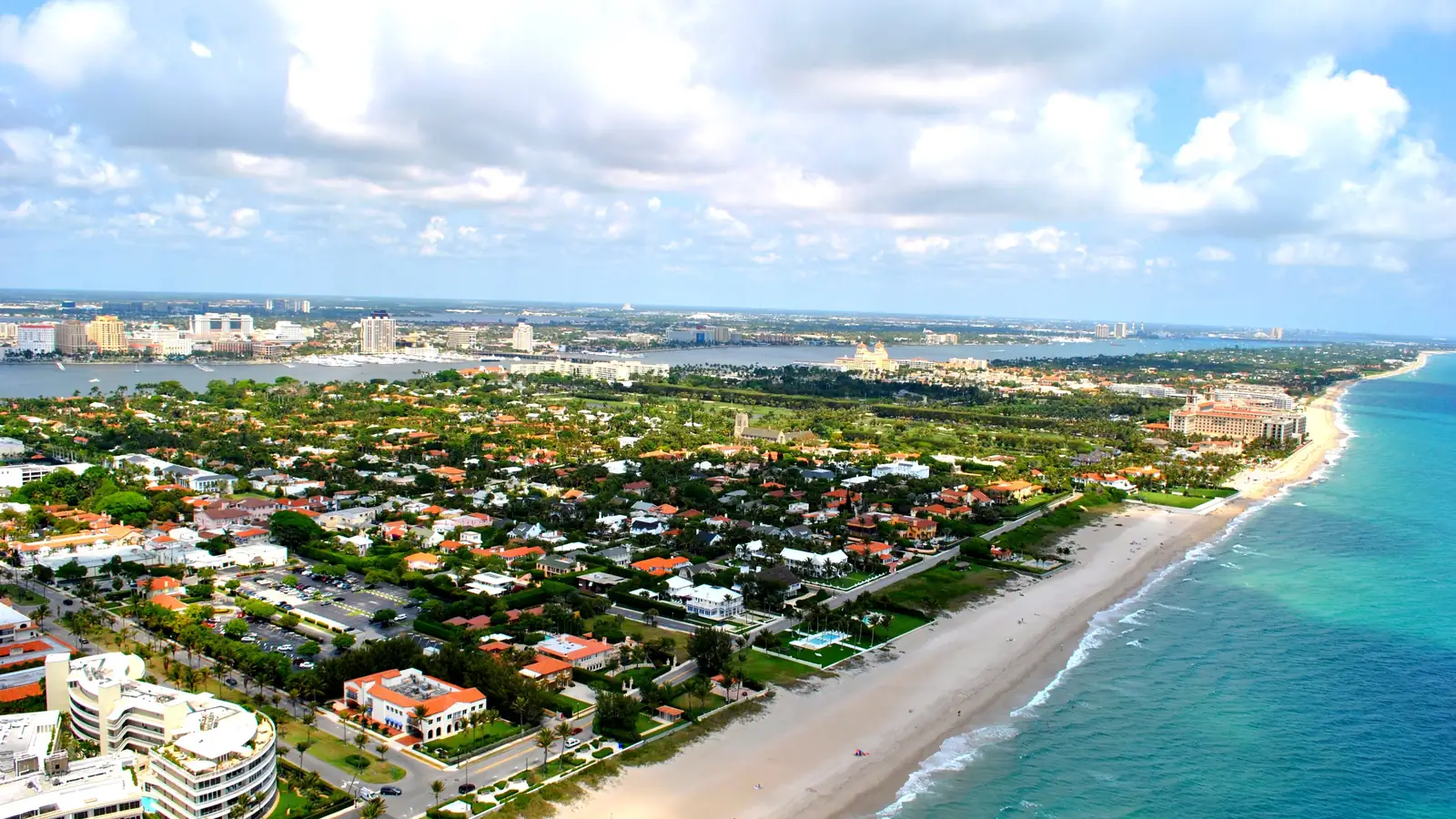 Bright aerial view over the island: red-roofed houses and green streets running to a long sand beach and turquoise surf on the right, a large hotel with twin towers at the centre right, and the lagoon and mainland skyline on the horizon under cumulus clouds.
