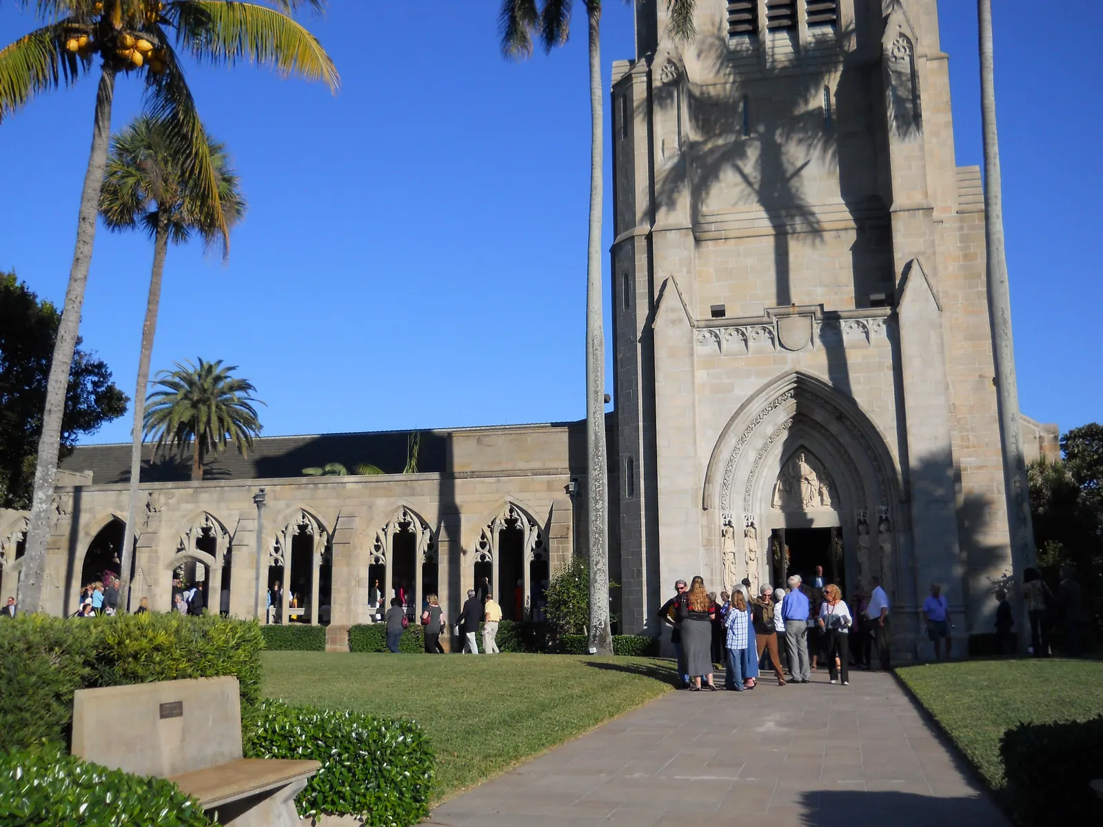 The tall Gothic stone tower and pointed-arch doorway of a church beside a cloister of open Gothic arches, with palms, a lawn and a stone bench, and a group of about twenty people gathered on the path by the door under a deep blue sky.
