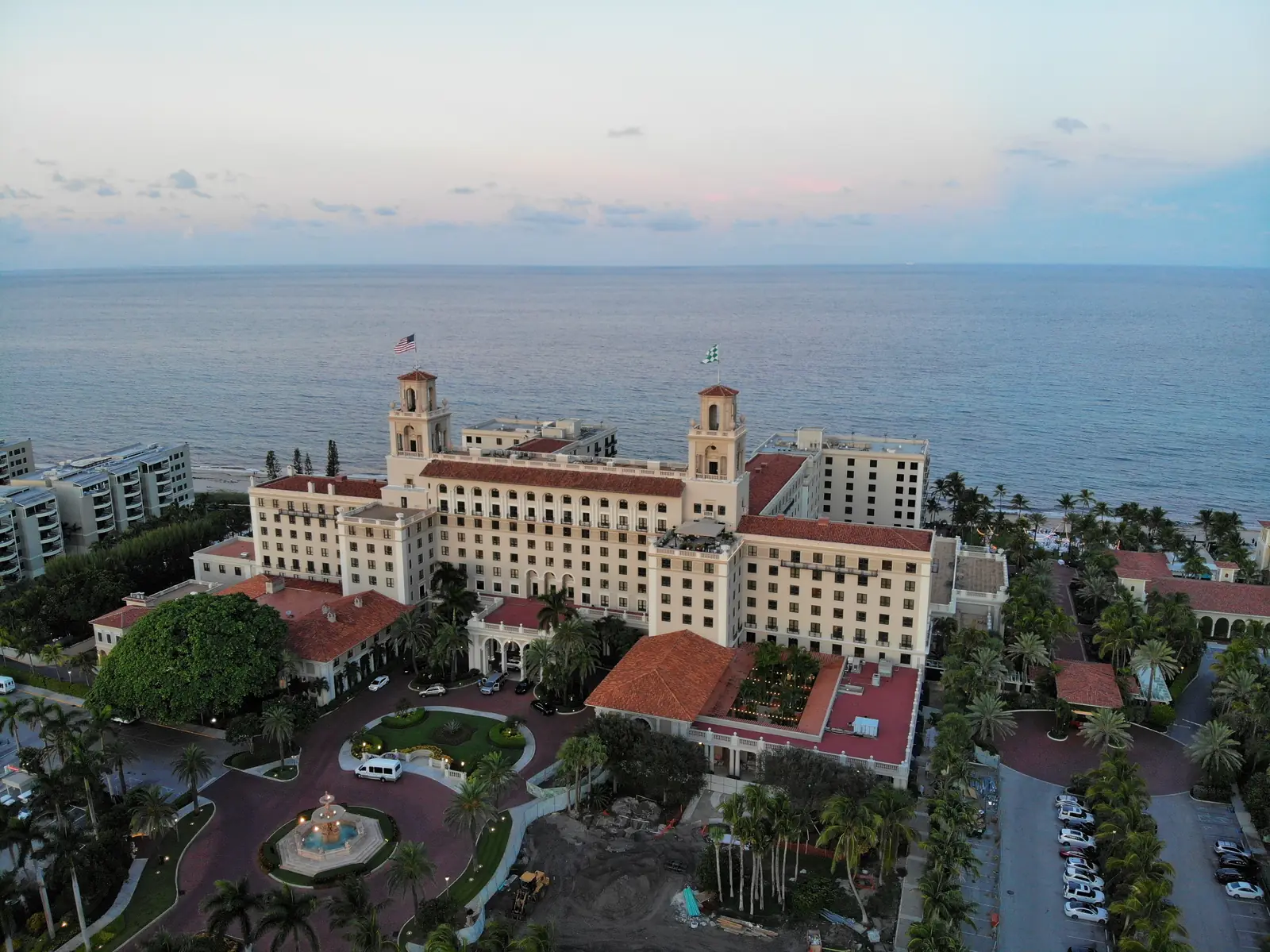 Elevated dusk view of a large cream hotel with twin towers and red tile roofs facing the Atlantic, with a circular fountain drive, palms, gardens and a parking area in the foreground and the calm ocean and a pale pink sky behind.