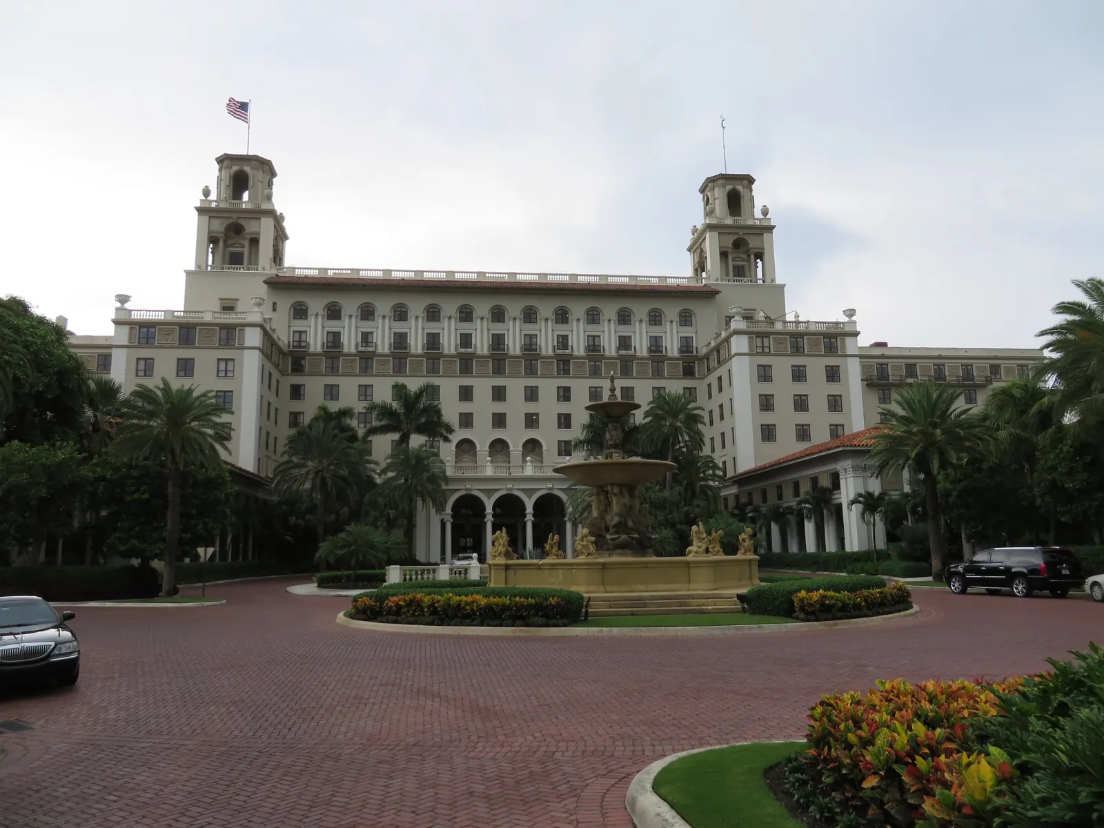 The front of a large cream hotel with twin towers, a flag and rows of arched windows, behind a tiered stone fountain in a round flower bed at the centre of a brick-paved drive, with palms and two dark cars at the edges, under an overcast sky.