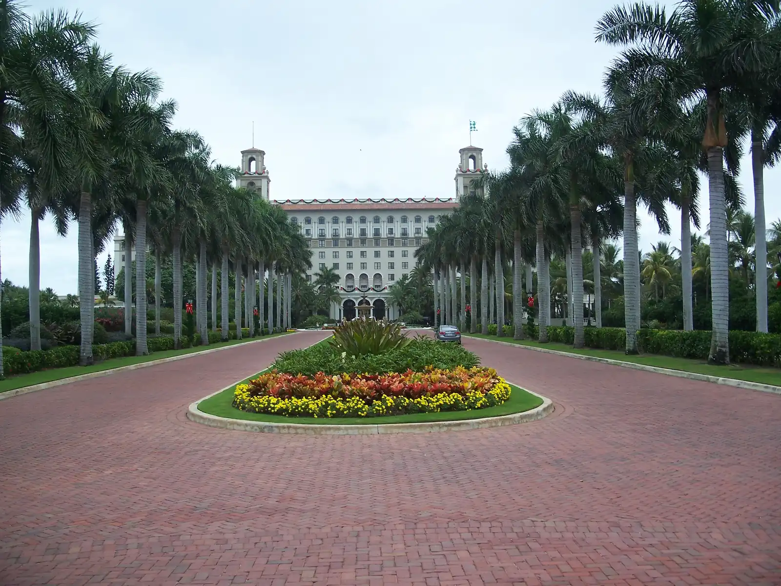 A long brick-paved drive lined on both sides by rows of tall palms, with a flower bed of red, orange and yellow blooms in the foreground, leading to a large cream hotel with twin towers at the far end.