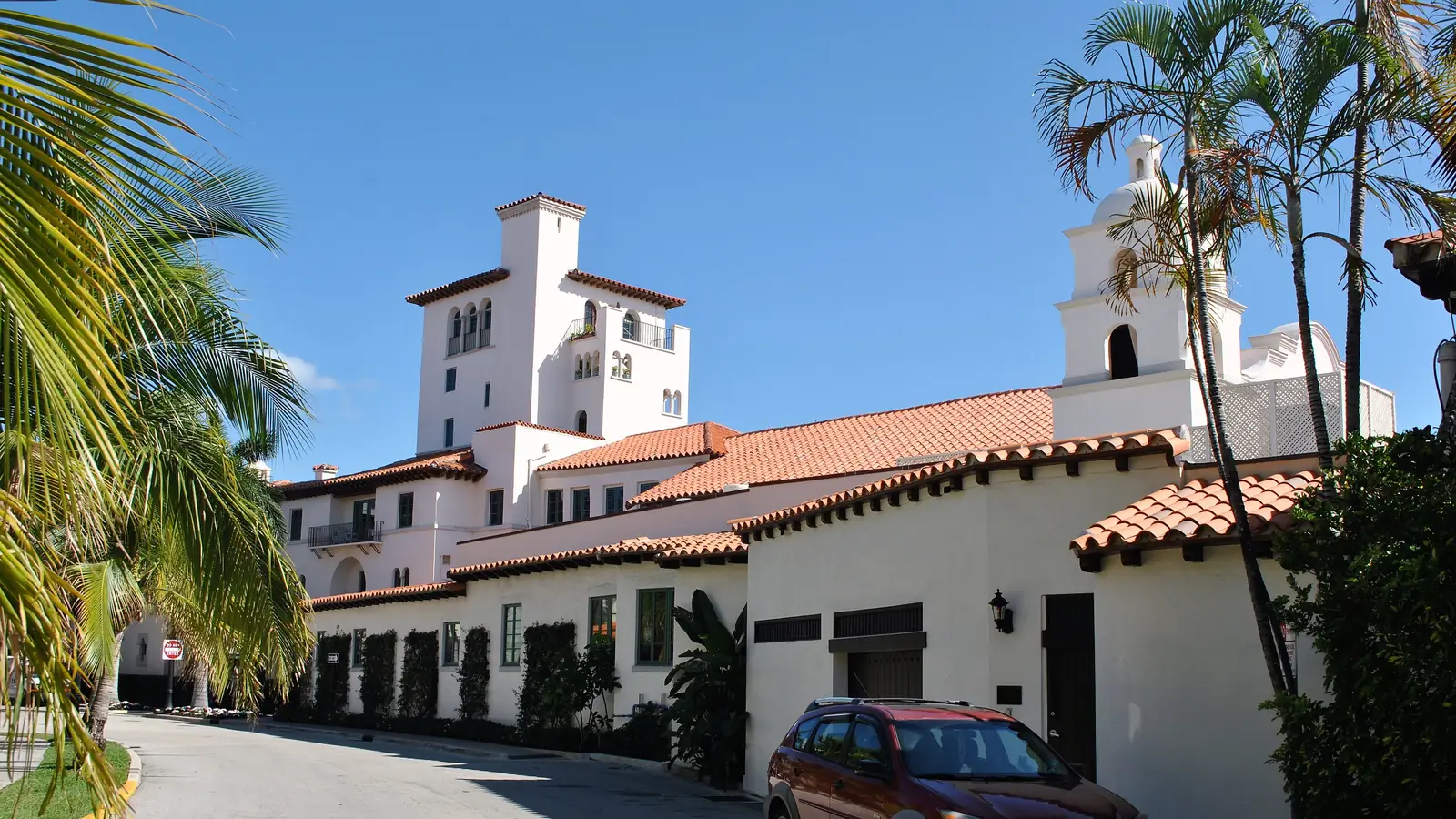 A white stucco Mediterranean-style complex with red tile roofs, a tall square tower and a small bell tower, along a curving driveway with palms, and a parked red hatchback in the foreground under a clear blue sky.