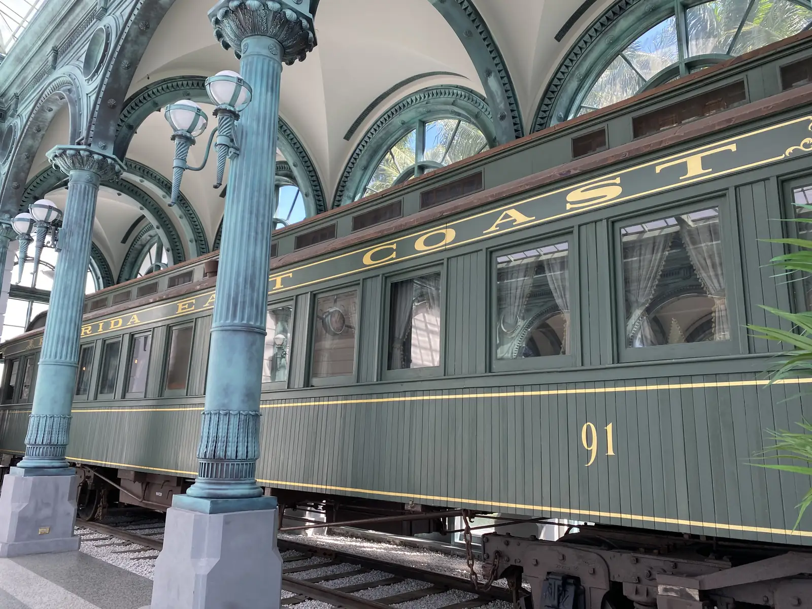 A dark green wooden railway passenger car lettered 'Florida East Coast' in gold with the number 91, under a pavilion of arched cast-iron columns painted pale green, lamp fixtures and tall arched windows.