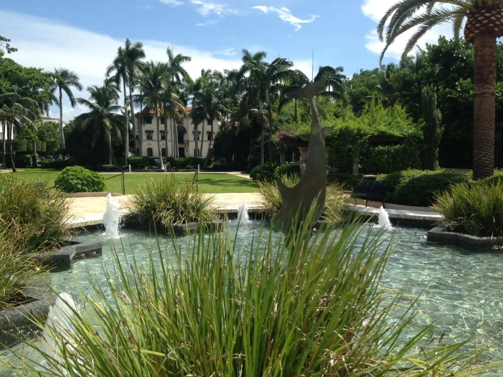 A shallow garden pool with small fountain jets seen through tall grasses in the foreground, a dark sculpture standing in the water, a lawn, palms and a cream building in the background.
