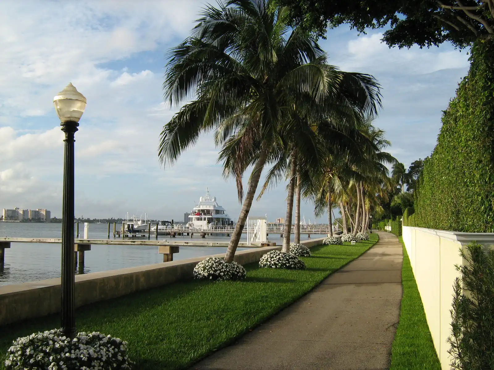 A paved path curving along a tall clipped hedge and white wall, with a lawn, white flowering shrubs, leaning coconut palms and an old-style lamp post on the water side, a seawall and docks with a large white yacht on the lagoon.
