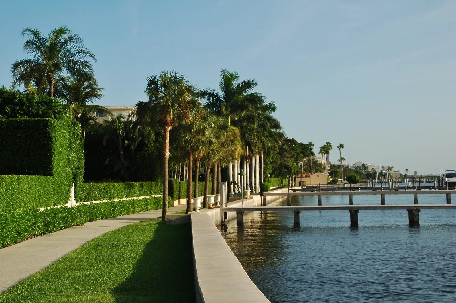A narrow walk on a grass strip between tall clipped hedges and a low seawall, with palms in low warm sunlight, wooden docks reaching into calm blue water and the far shore of the lagoon on the right.