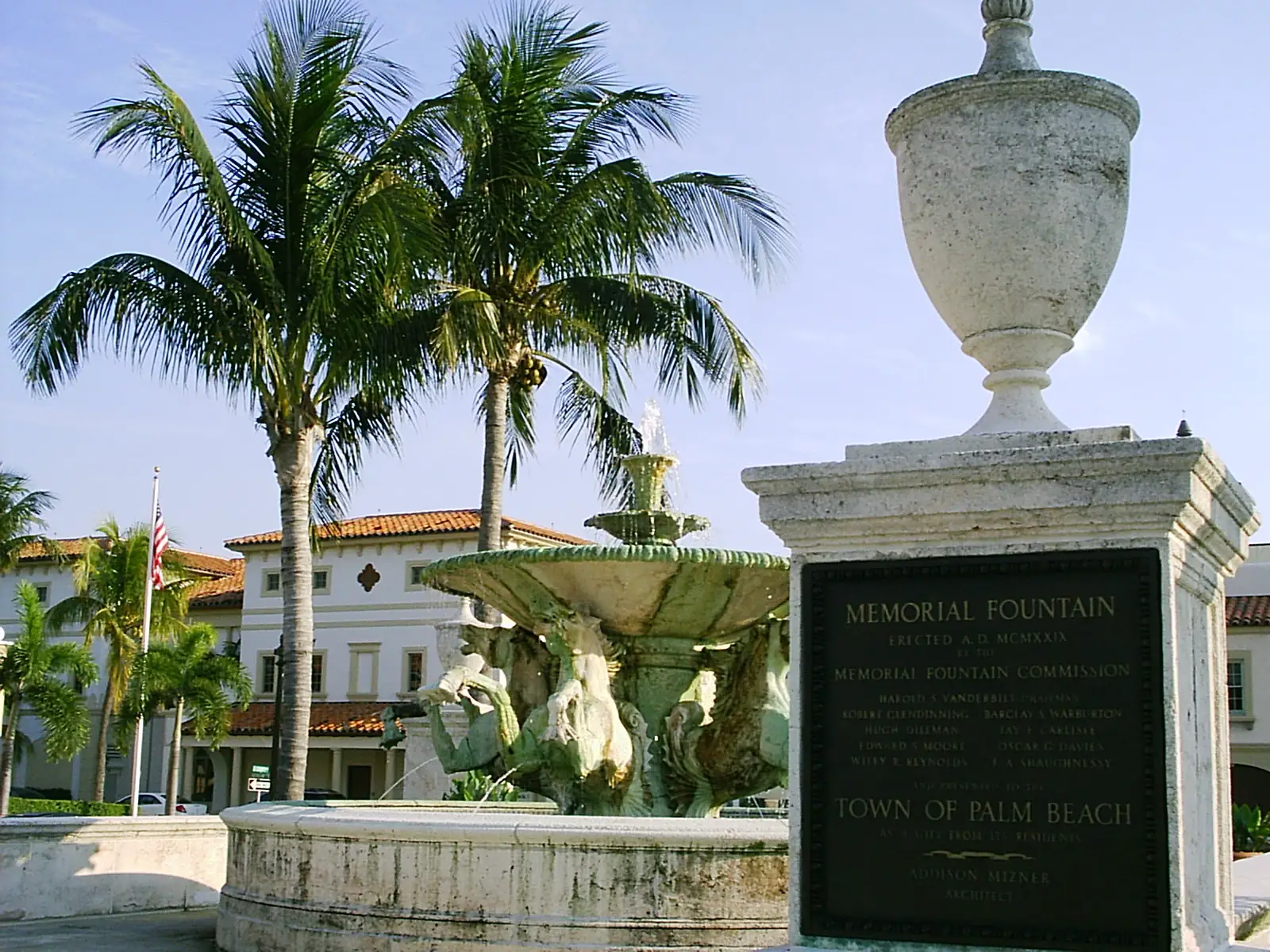 A weathered stone fountain with sculpted figures and a tiered basin beside a stone pedestal topped by an urn, whose bronze plaque reads 'Memorial Fountain' and 'Town of Palm Beach', with palms and a cream building with a red tile roof behind.