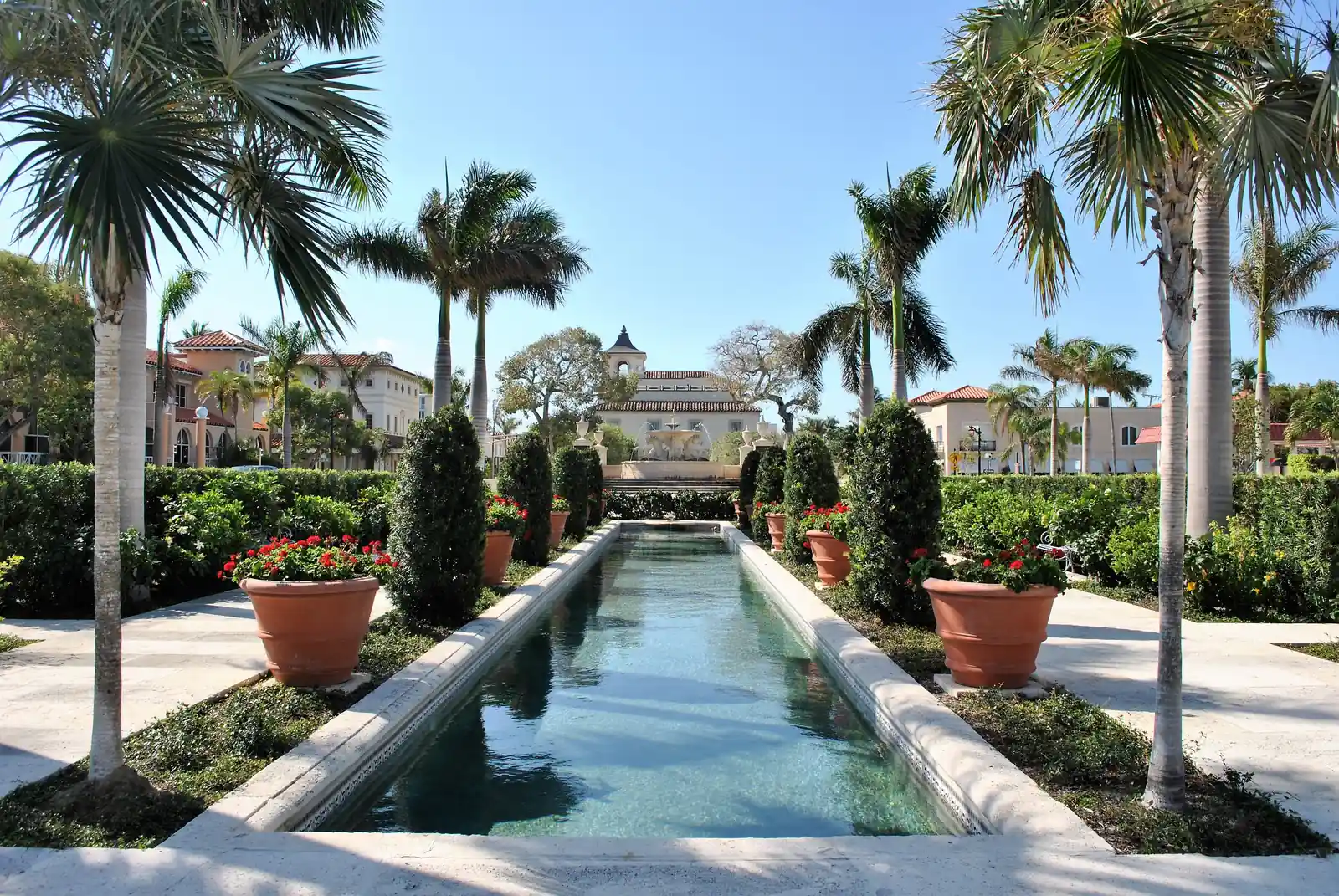 A long rectangular reflecting pool flanked by clipped columnar shrubs, terracotta pots of red geraniums and tall palms, leading the eye to cream buildings and a small domed tower beyond, under a clear sky.