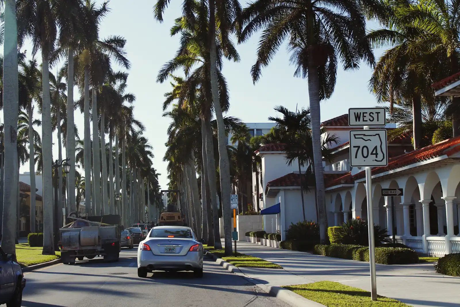 A street lined with very tall royal palms, a silver sedan and a work truck heading away, a 'West 704' route sign at right beside a white building with arched arcades and red tile roofs.