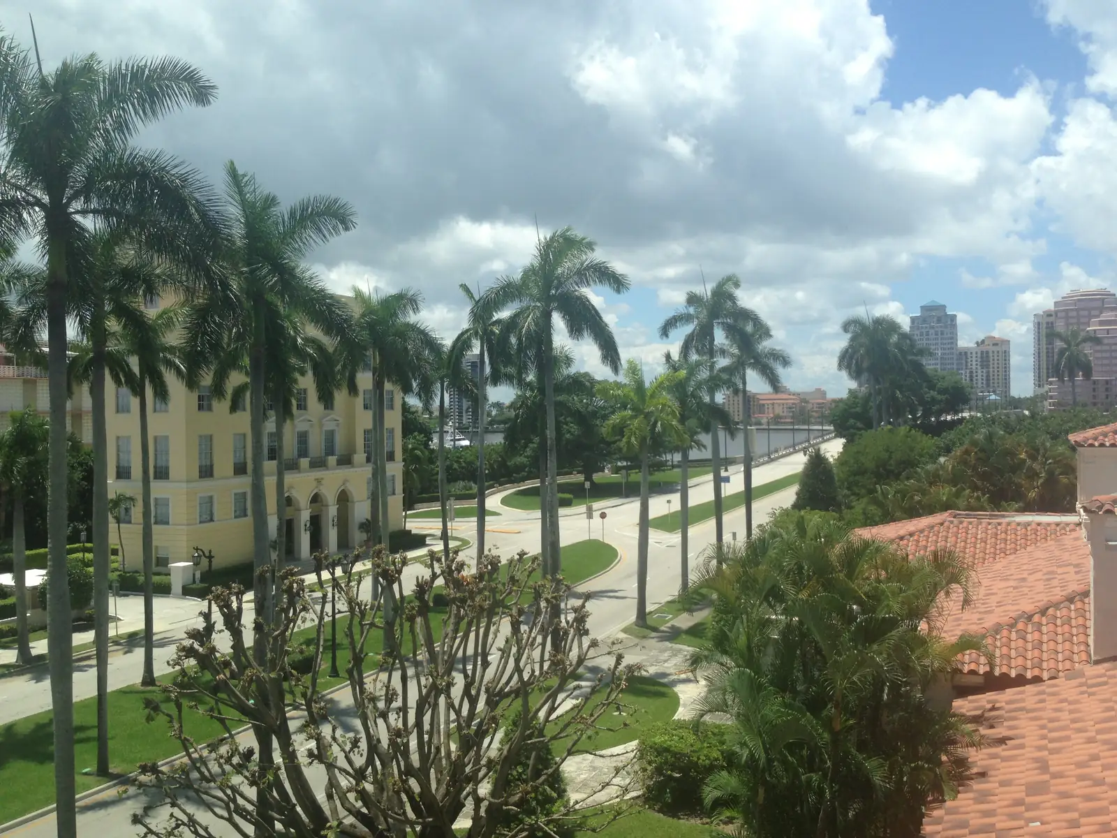 Elevated view over red tile roofs and palms to a curving palm-lined road and a yellow multi-storey building at left, with the lagoon and mainland towers on the horizon under heavy white clouds.