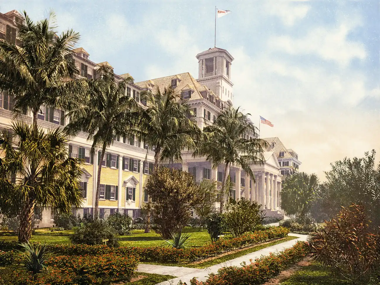 A hand-coloured photochrom of a very large yellow wooden hotel with green shutters, a white columned portico and a square cupola flying flags, set behind palms, flower beds and a curving garden path.