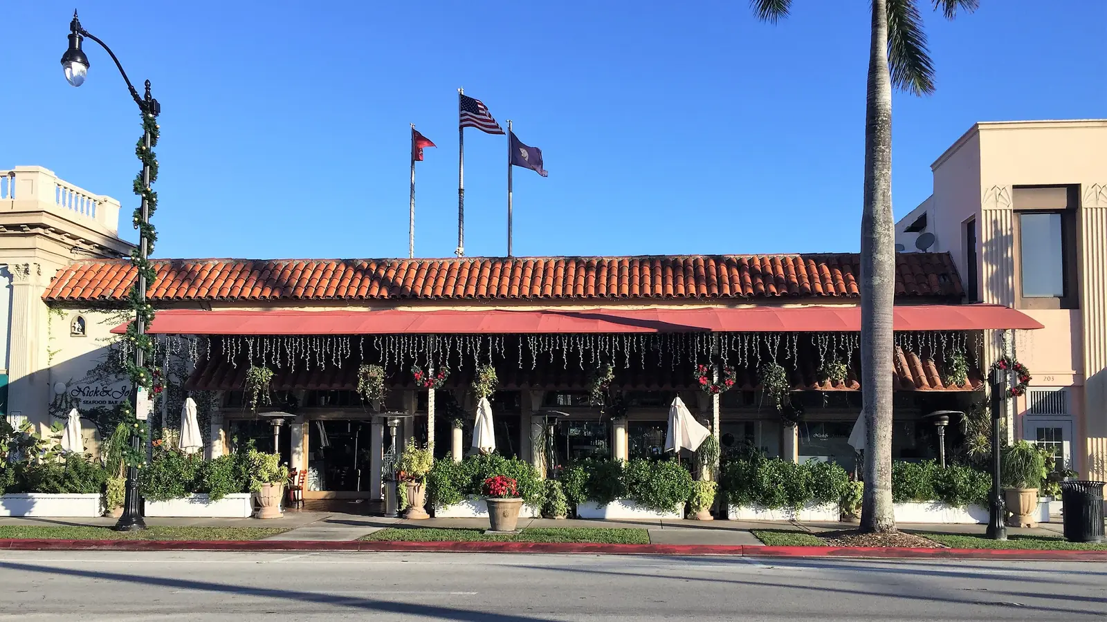 A low building with a red tile roof and a long red awning hung with string lights and holiday wreaths, potted plants and folded patio umbrellas along its front, three flags on poles above, seen across an empty street with a painted parking space number.