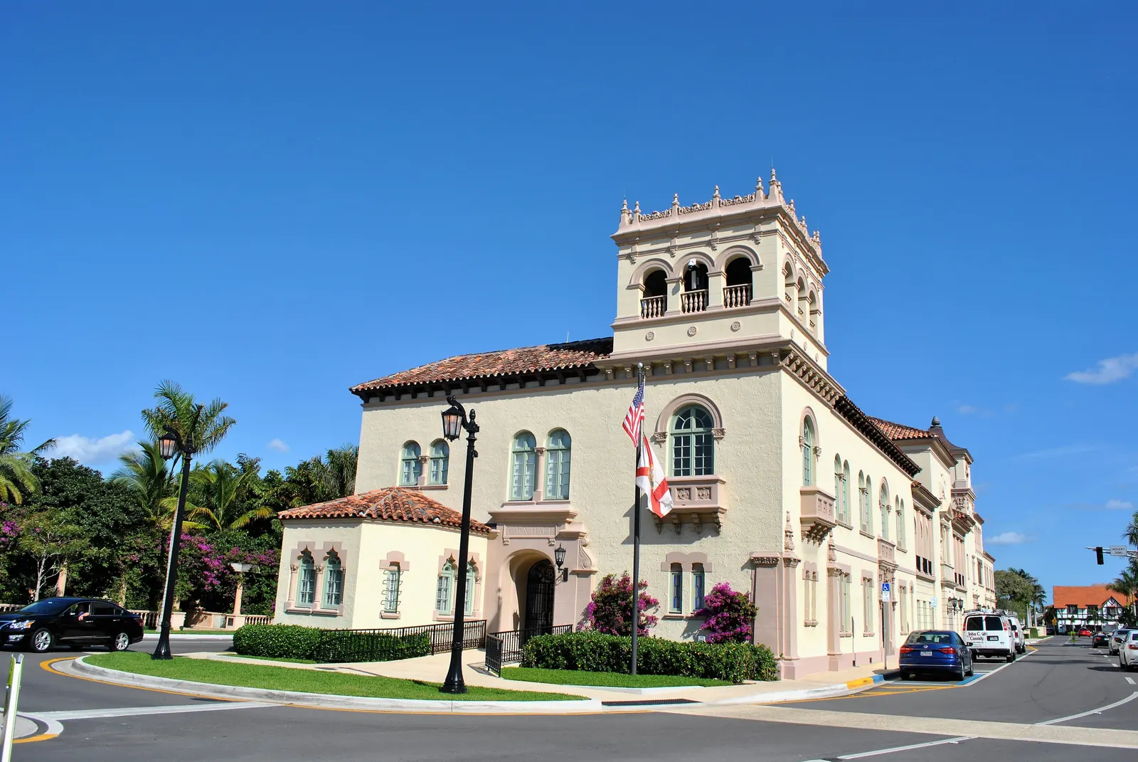 A cream Mediterranean Revival building with a tall arcaded tower, red tile roofs and arched windows, flying the United States and Florida flags, on a street corner with palms, lamp posts, bougainvillea and a few parked cars under a clear blue sky.