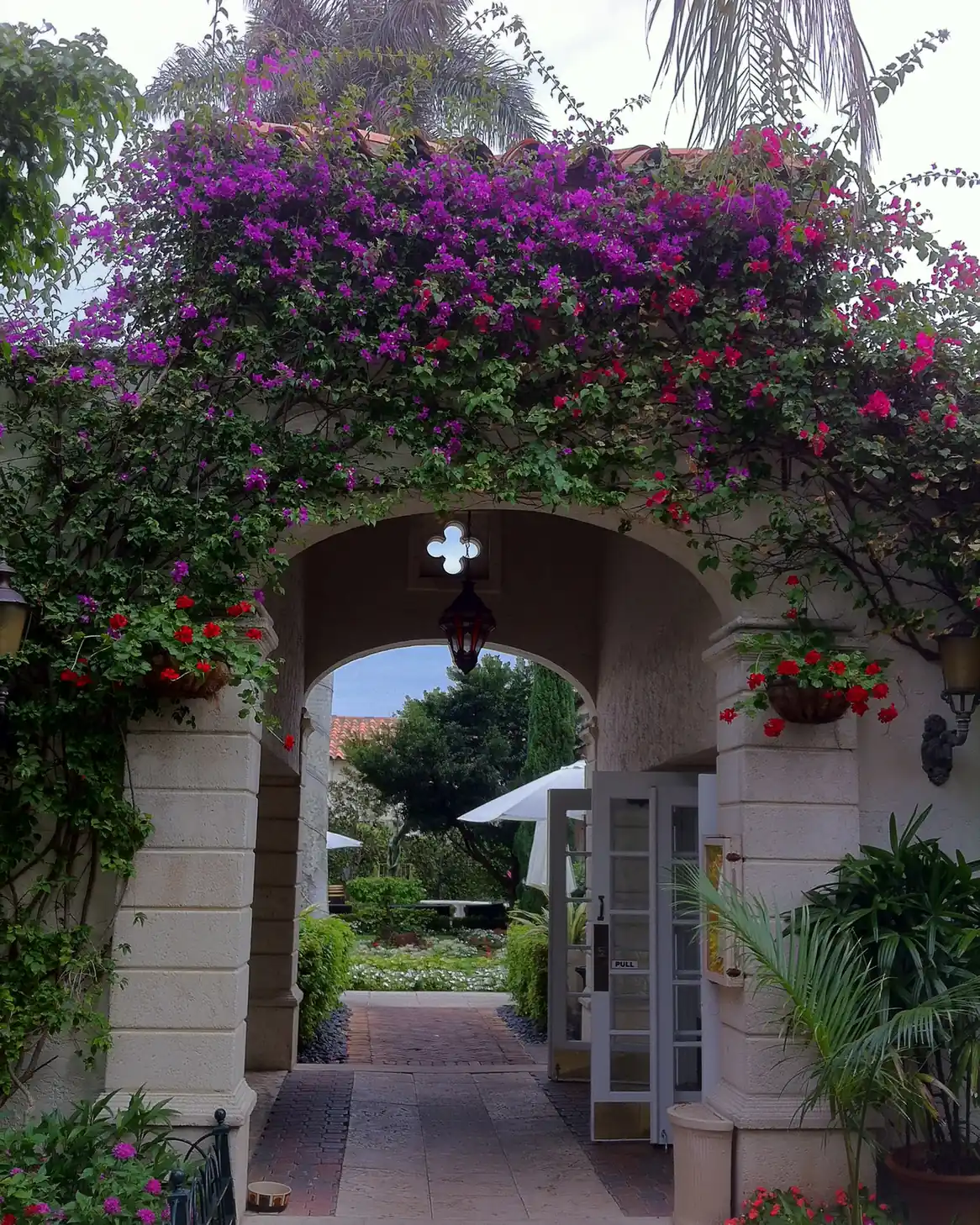 A stucco archway draped in magenta bougainvillea with hanging baskets of red geraniums, opening onto a brick-paved passage that leads to a sunny garden with white umbrellas.