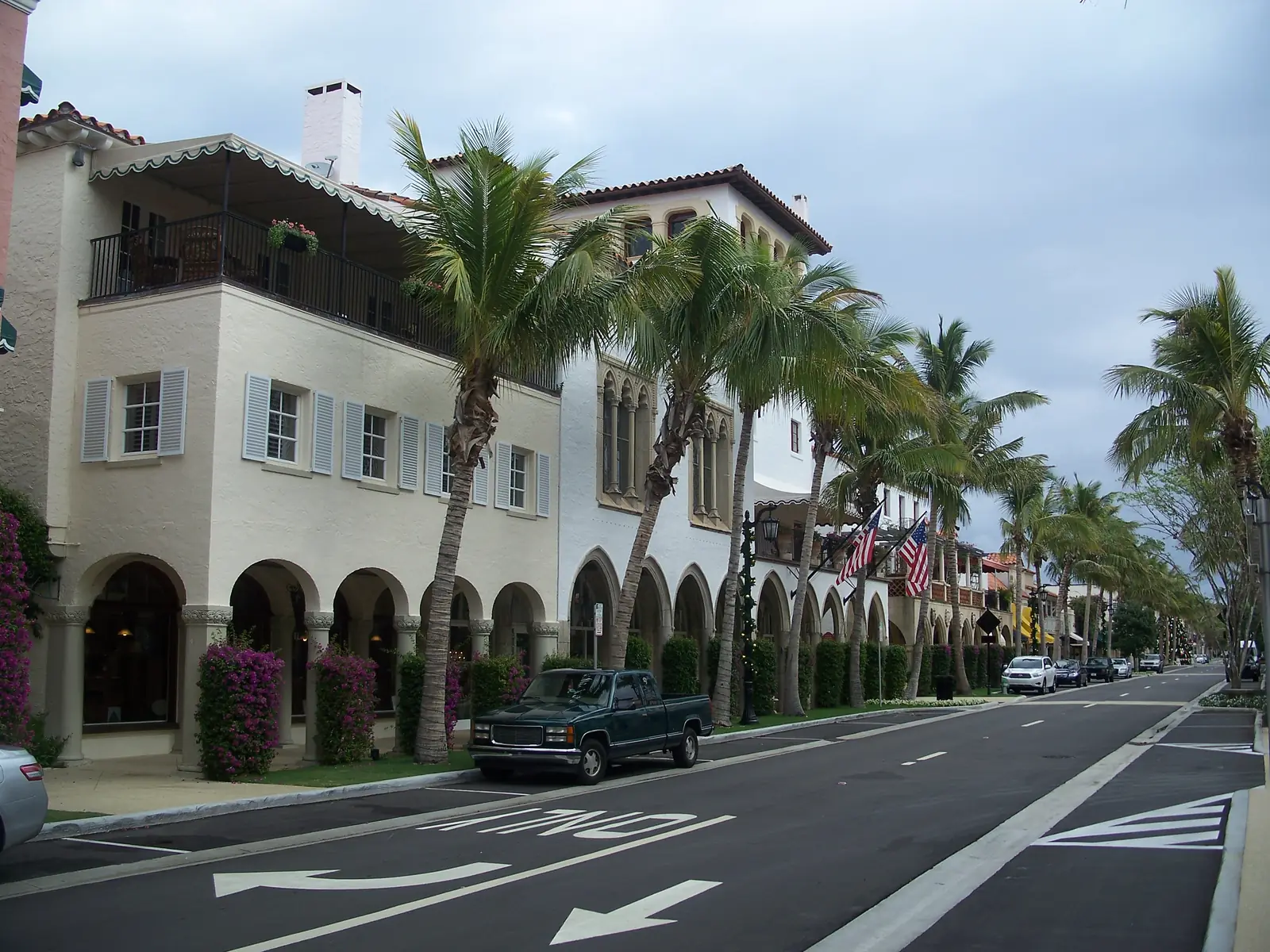 A white Mediterranean-style building with a balcony, shuttered windows and a long arcade of round arches along the street, palms and bougainvillea in front, American flags further down, and an empty two-lane street with painted arrows under a grey sky.