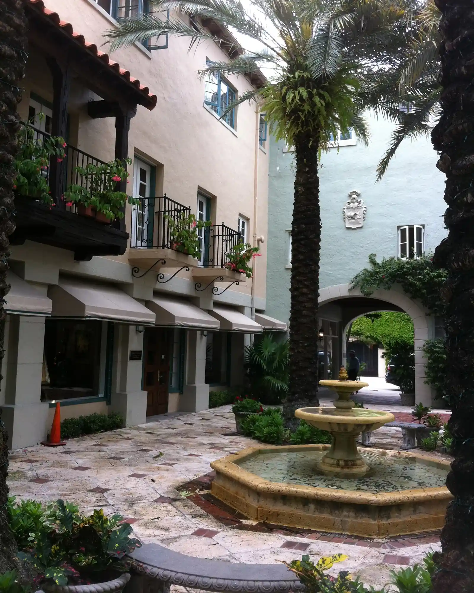 A narrow courtyard passage with a tiered stone fountain on a patterned stone floor, a palm, potted plants, wrought-iron balconies and awnings on the buildings, and an archway at the far end.