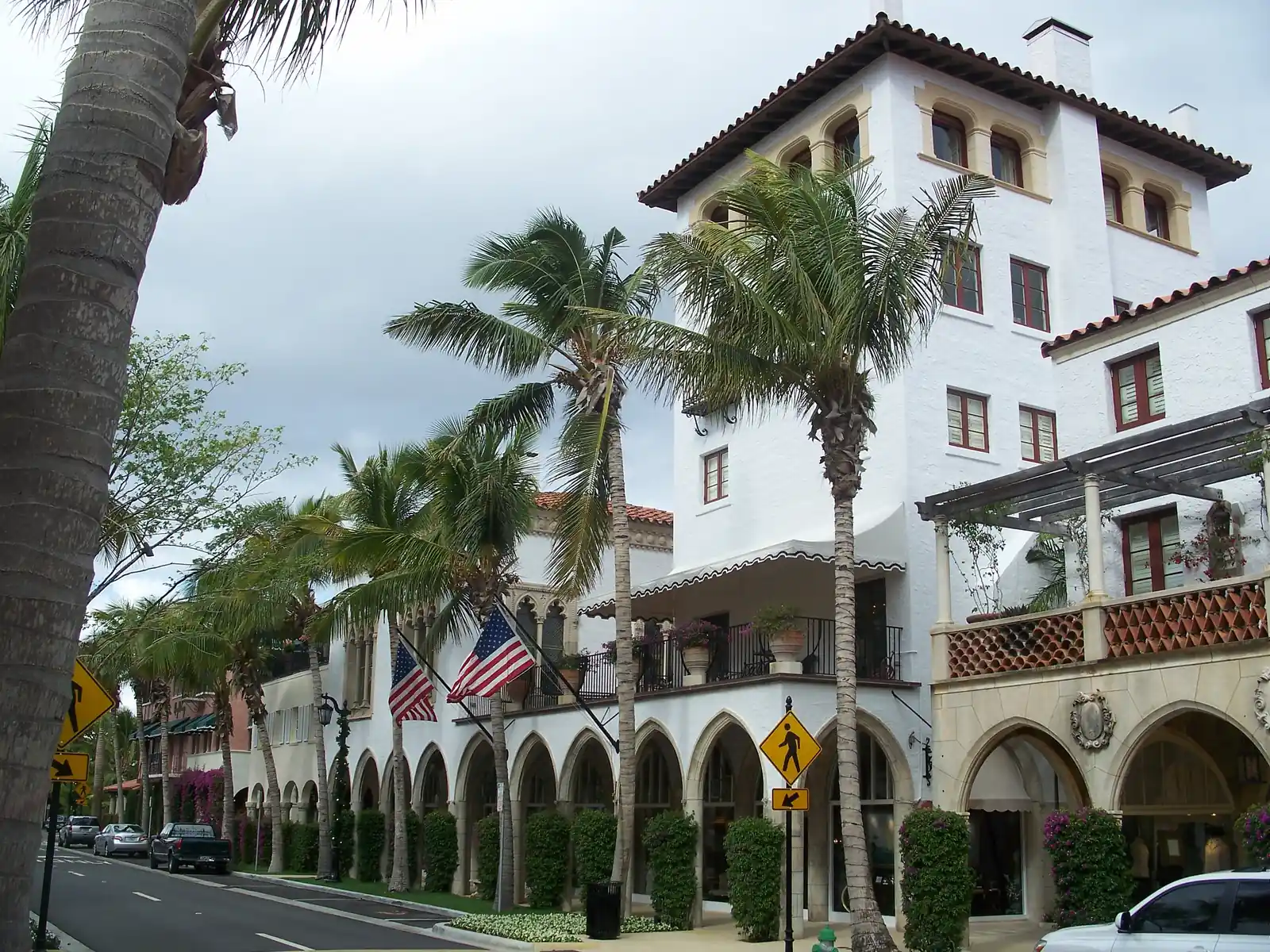 The tall white tower of a Mediterranean-style building with a red tile roof, pointed-arch arcades at street level, wrought-iron balconies, American flags, palms and pink bougainvillea, seen from the sidewalk under a cloudy sky.