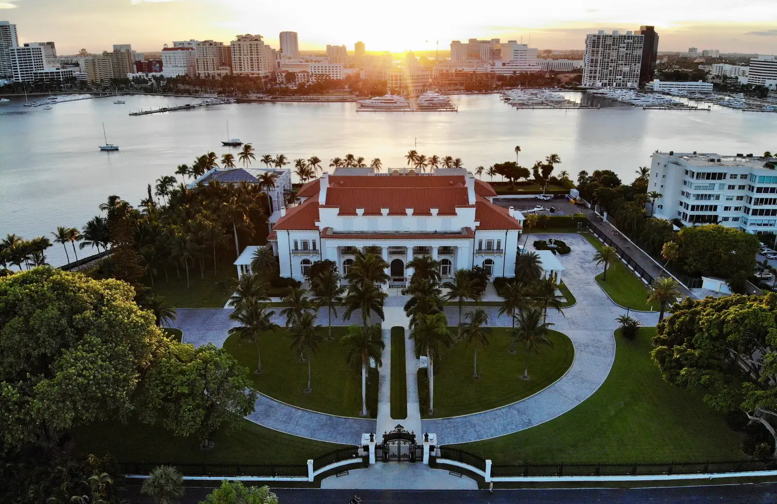 Elevated view at sunset over a white mansion with red tile roofs set in a palm-lined lawn with a curving drive and front gate, the lagoon behind it with boats and docks, and the mainland skyline across the water with the low sun flaring between towers.