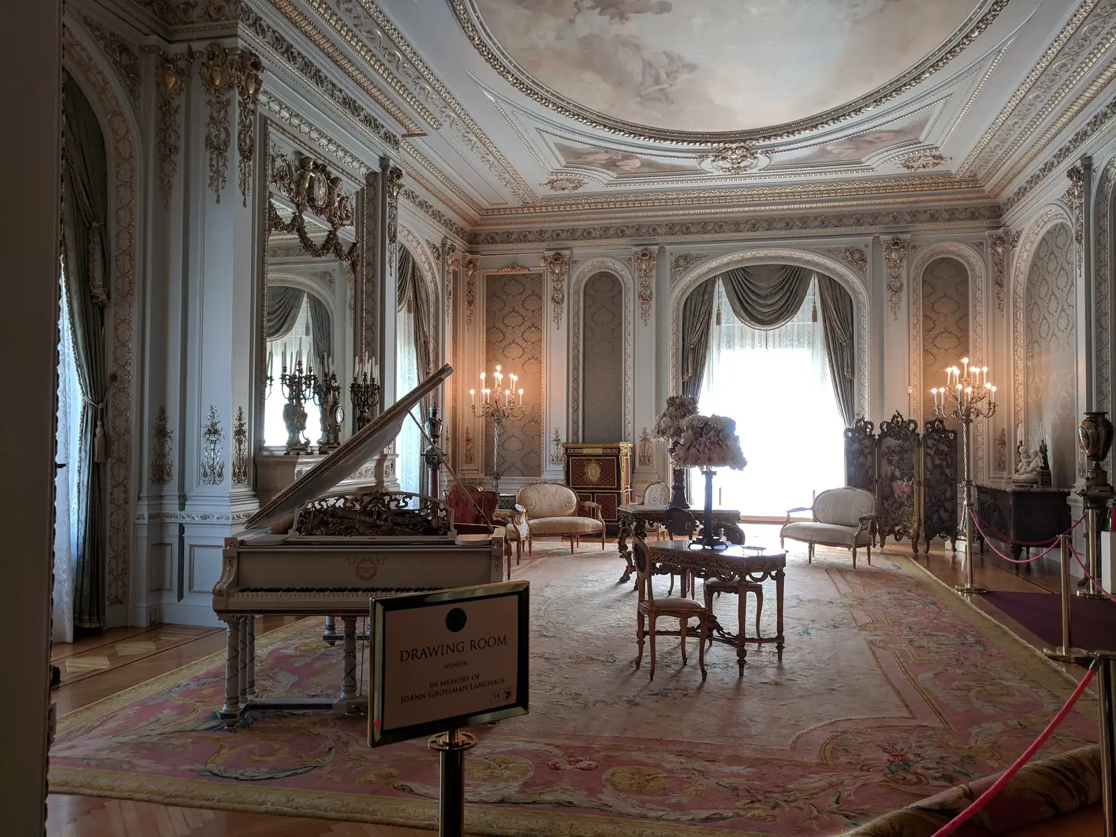 An ornate white and gold drawing room with a painted oval ceiling, mirrored panels, candle sconces and draped windows, a gilded grand piano, period chairs and tables on a large pink rug, roped off with a small sign reading 'Drawing Room'.