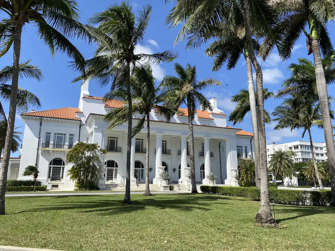 The white neoclassical front of a large mansion with a row of tall columns, a red tile roof and arched windows, seen across a sunny lawn through tall leaning coconut palms under a blue sky with light cloud.