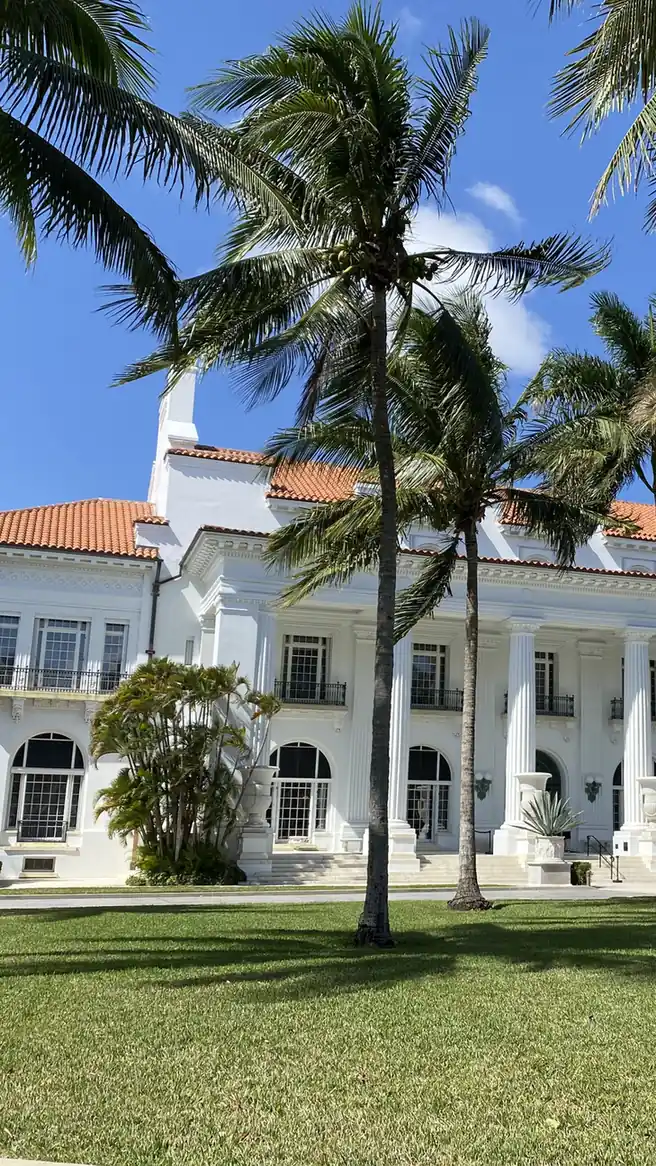 The white neoclassical front of a large mansion with a row of tall columns, a red tile roof and arched windows, seen across a sunny lawn through tall leaning coconut palms under a blue sky with light cloud.