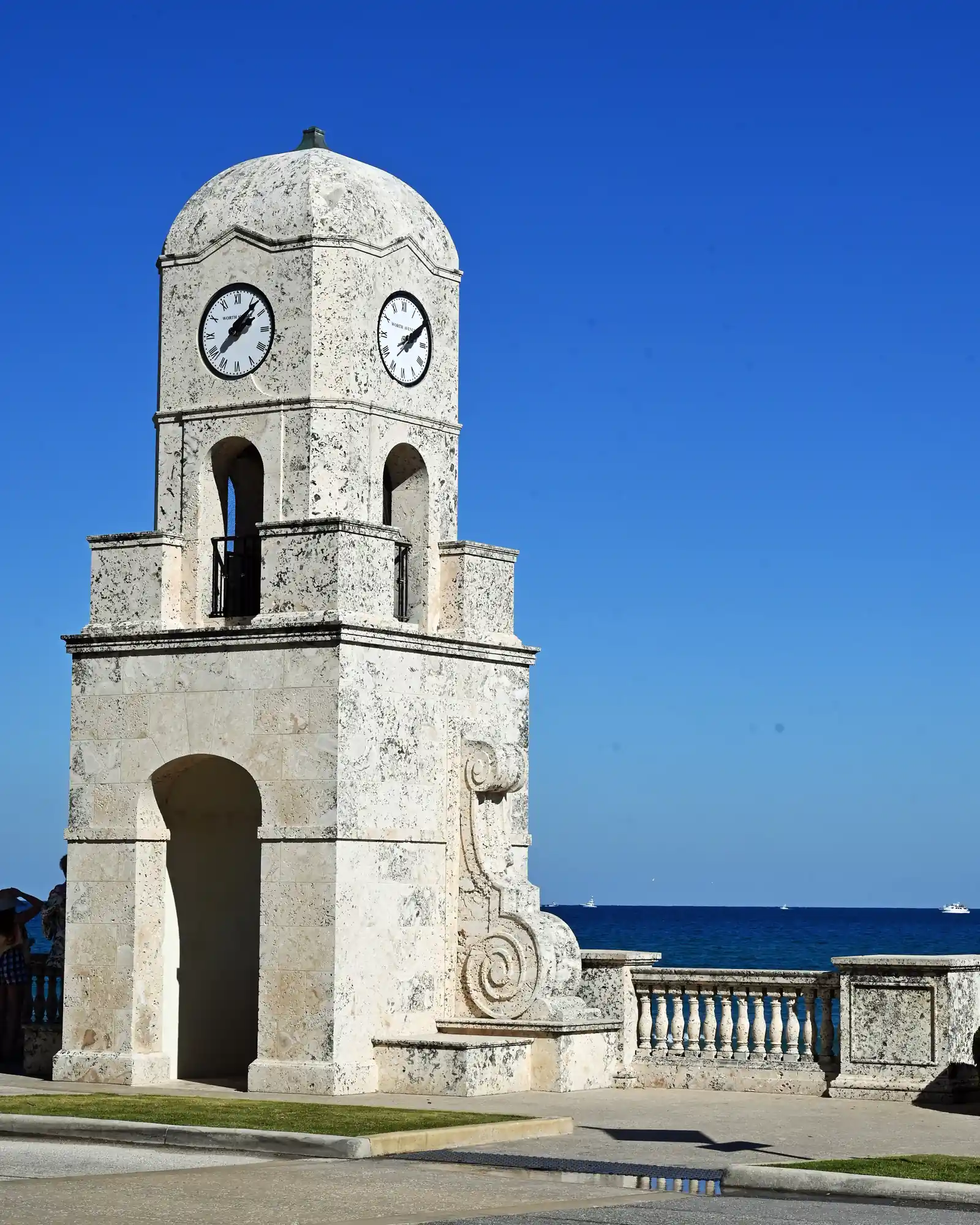 A coral-stone clock tower with a domed top, clock faces on two sides and an open arched base, against a deep blue sky, with a balustraded seawall and the blue Atlantic behind it.
