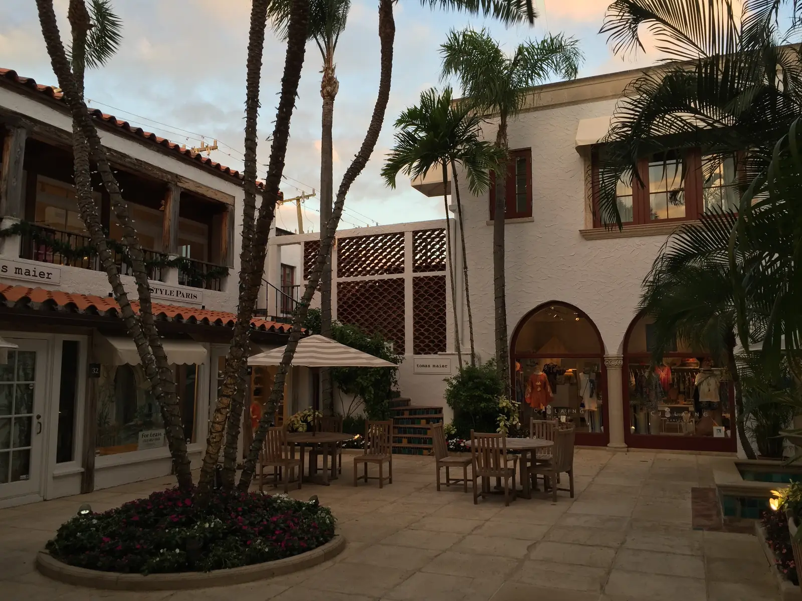 A paved courtyard ringed by two-storey stucco shops with arched windows and red tile roofs, a round planter of palms and flowers in the centre, wooden café tables and chairs under a striped umbrella, in soft evening light.