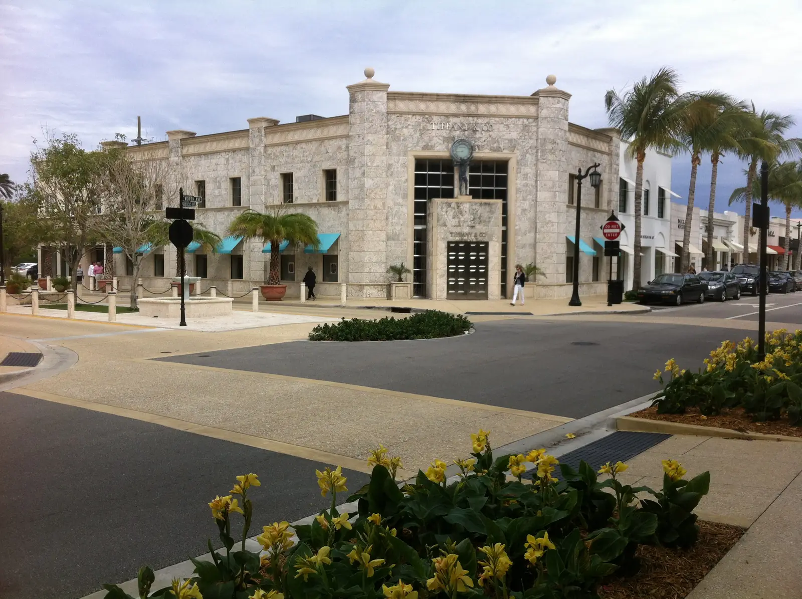 A pale coral-stone corner building with a tall framed window bay over its entrance and teal awnings, seen across a curving intersection; yellow canna flowers in the foreground bed, palms and white shopfronts continuing down the street, a few pedestrians small in the distance.