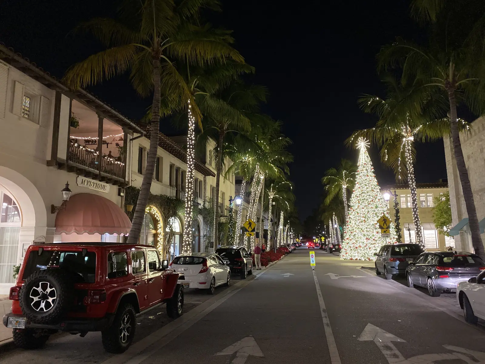 Night view down Worth Avenue with its palms wrapped in white string lights, a tall lit holiday tree on the right side of the street, cars parked along both curbs and a red Jeep in the foreground, low arcaded shopfronts and a lit balcony on the left.