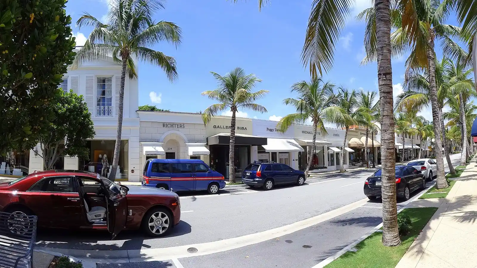 Wide sunny view along Worth Avenue: a red sedan with an open door and other parked cars, palms and a leafy tree along the curb, white two-storey shopfronts with dark awnings, a wide sidewalk on the right under a deep blue sky.